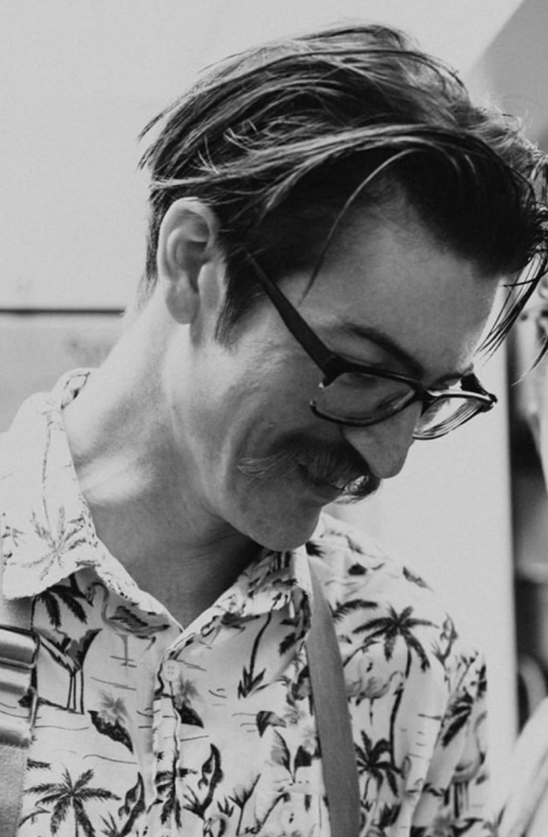 Nicholas Webber, looking down towards likely a plate of food that is not visible in this picture. There's a slight smile and he wears glasses and has long hair. His buttoned-up shirt has palm trees on it.